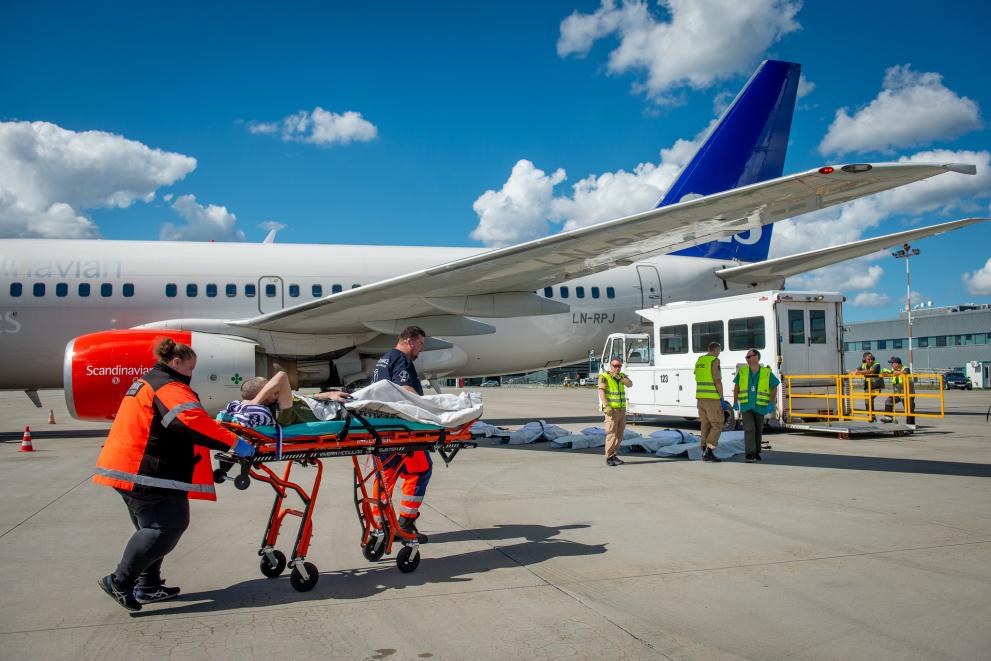 A patient on a brancard with 2 medics, in the back a plane awaiting the arrival of the patient.