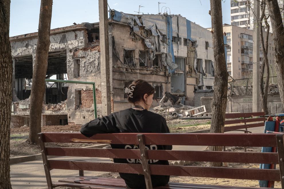 A woman sitting on a park bench seen from the back, in the background ruined buildings.